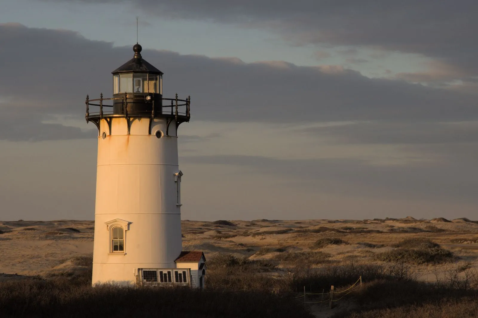 A white lighthouse standing in dune grass under a soft evening sky