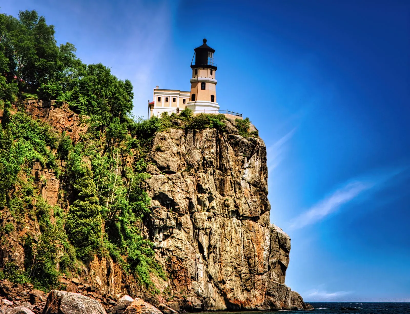 A lighthouse on top of a sheer rock cliff above a lake under a deep blue sky