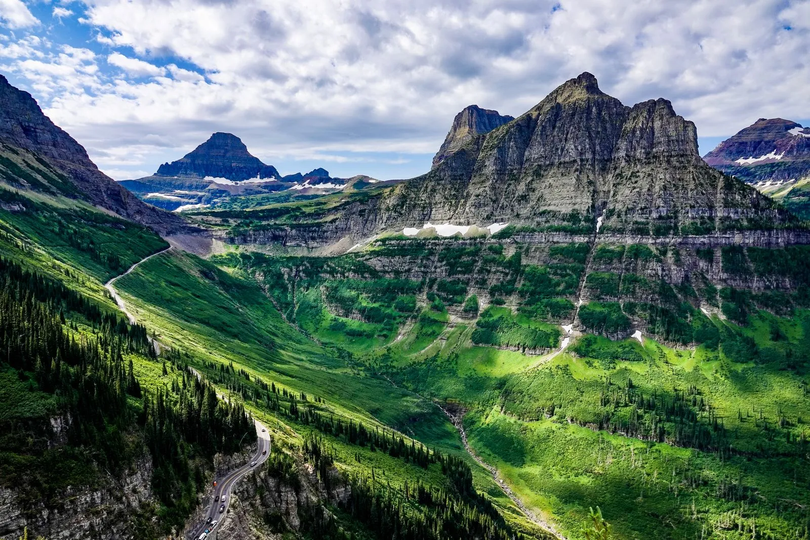 A green glacial valley below jagged gray peaks, a road winding up its side
