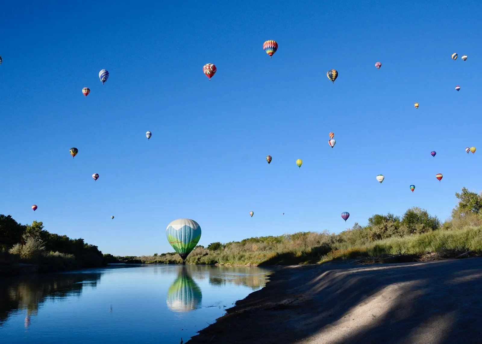Dozens of hot air balloons in a clear sky, one reflected in a still river below
