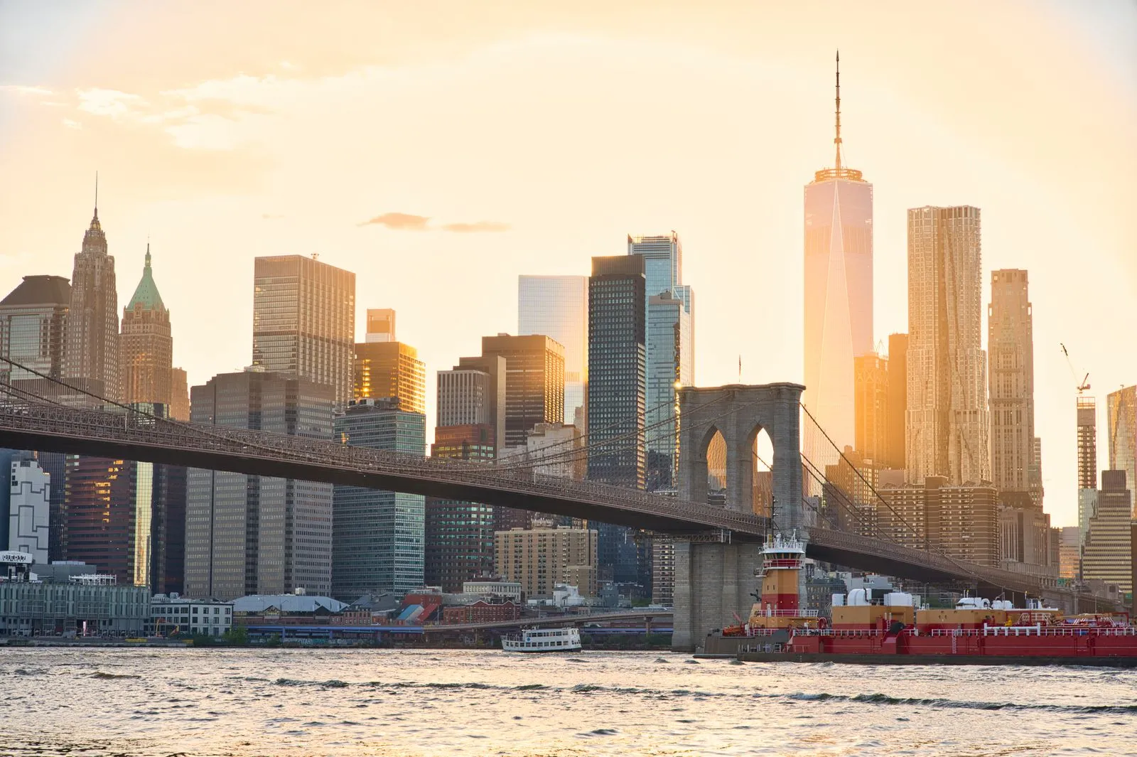 A stone suspension bridge crossing a river in front of a skyline of towers at sunset