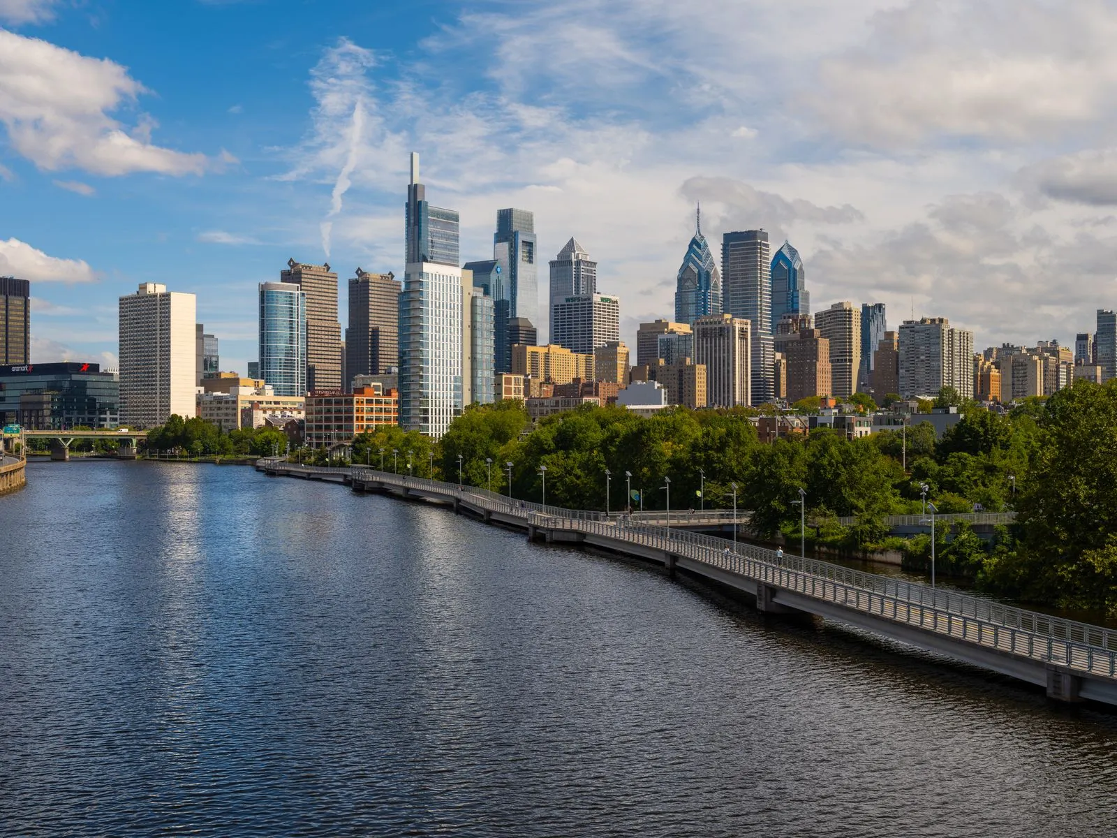 A river and riverside boardwalk leading toward a skyline of glass and stone towers