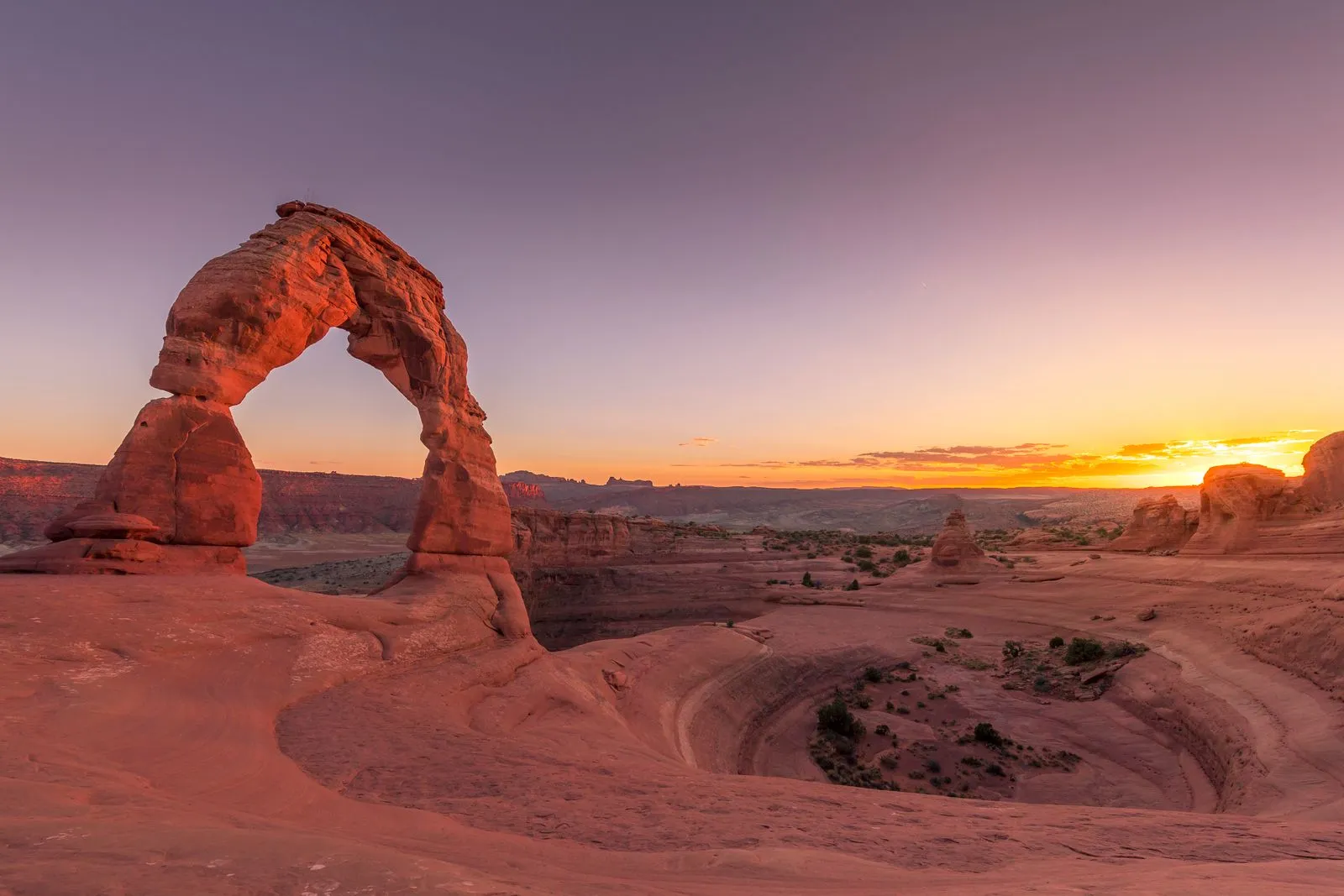 A lone red sandstone arch on a slickrock bowl at sunset