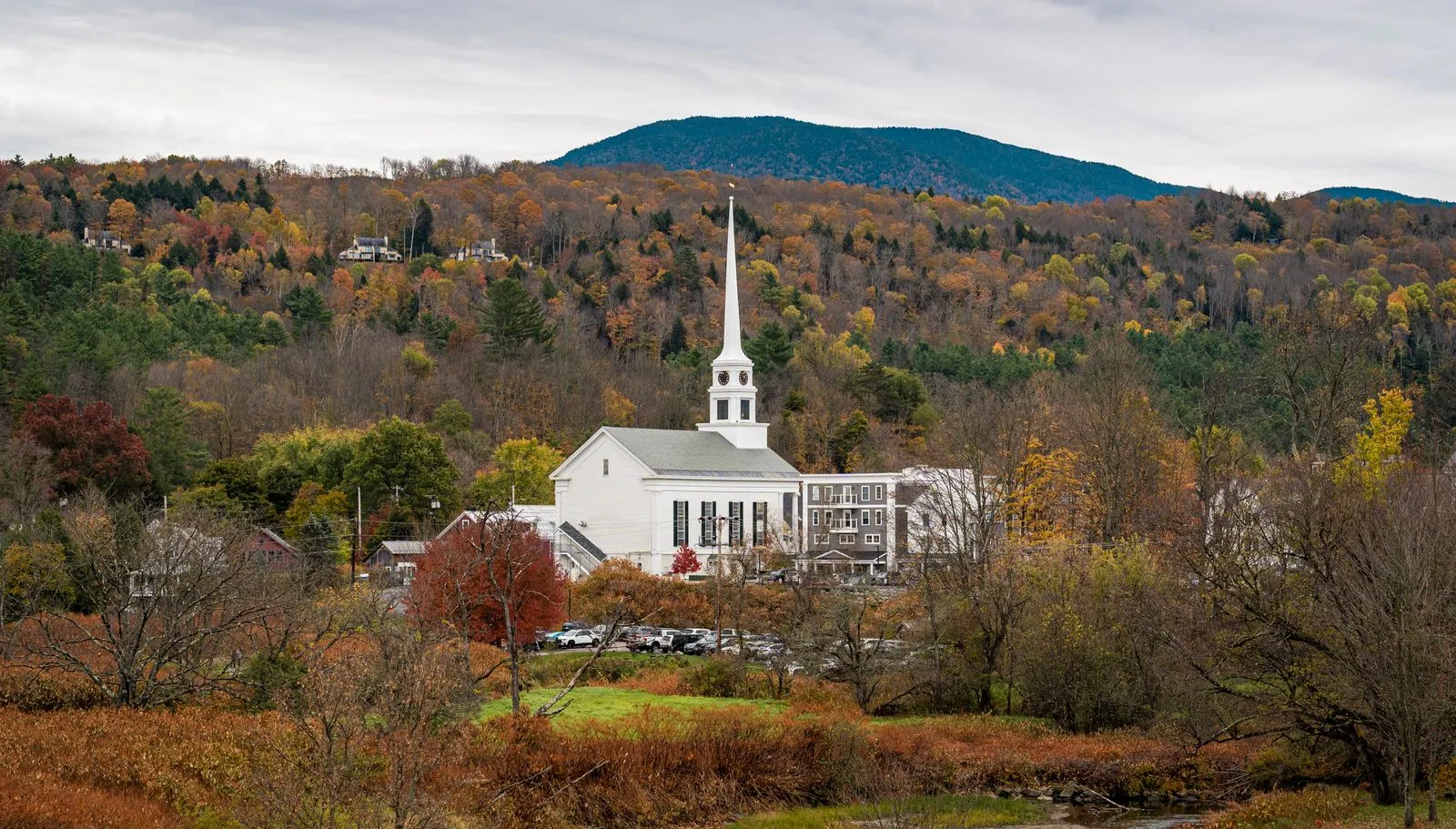 A white church with a tall steeple among autumn trees below a rounded mountain