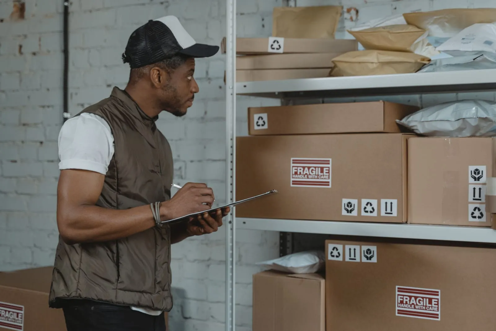 Warehouse worker in a trucker cap checks a clipboard beside shelves of cardboard boxes marked fragile and packages in mailers