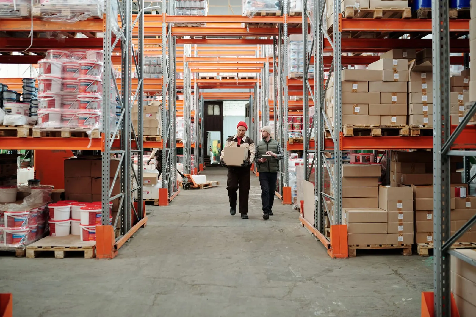 Two warehouse workers walk down a wide aisle between tall orange racks stacked with boxes and paint buckets, one carrying a carton